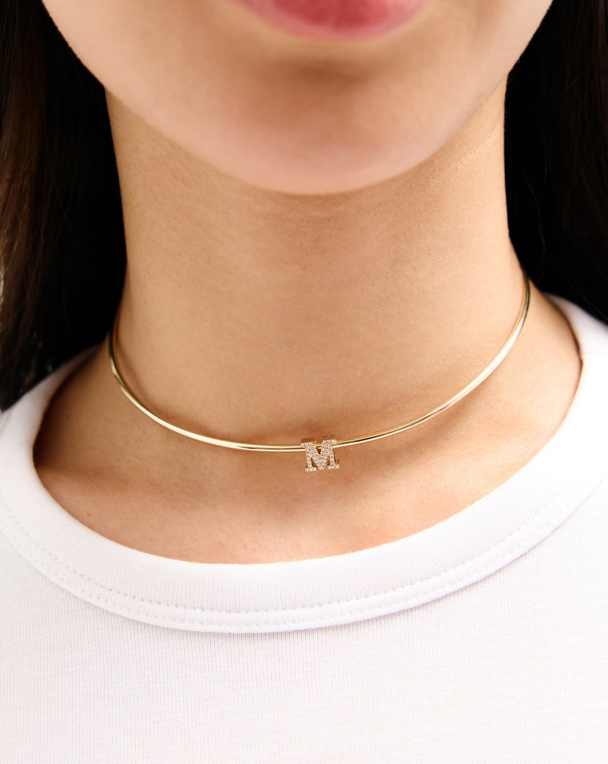 A close-up of a model's neck wearing a delicate gold bangle choker necklace with a small capital letter 'M' charm. global