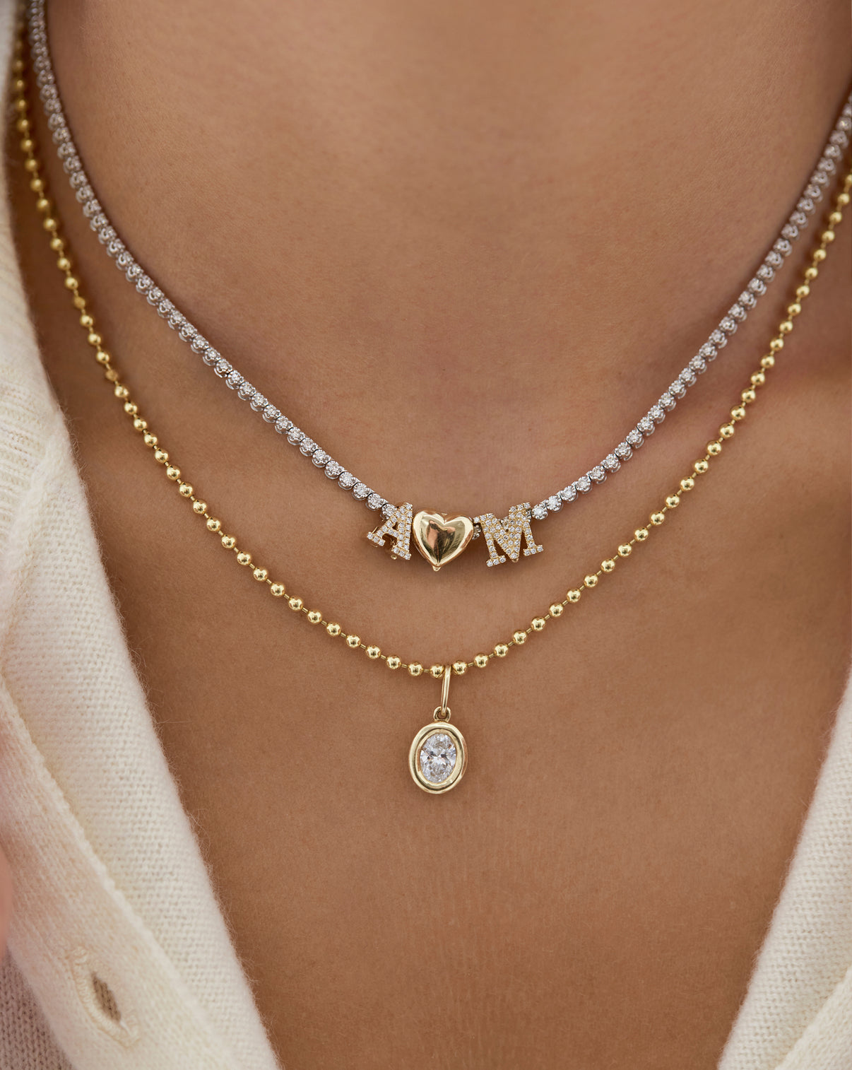 Close-up of a person wearing layered necklaces: a silver-tone beaded necklace with a heart-shaped charm outlined with small stones and the letters A and M, plus a gold-tone beaded necklace with a small oval stone globalpendant.