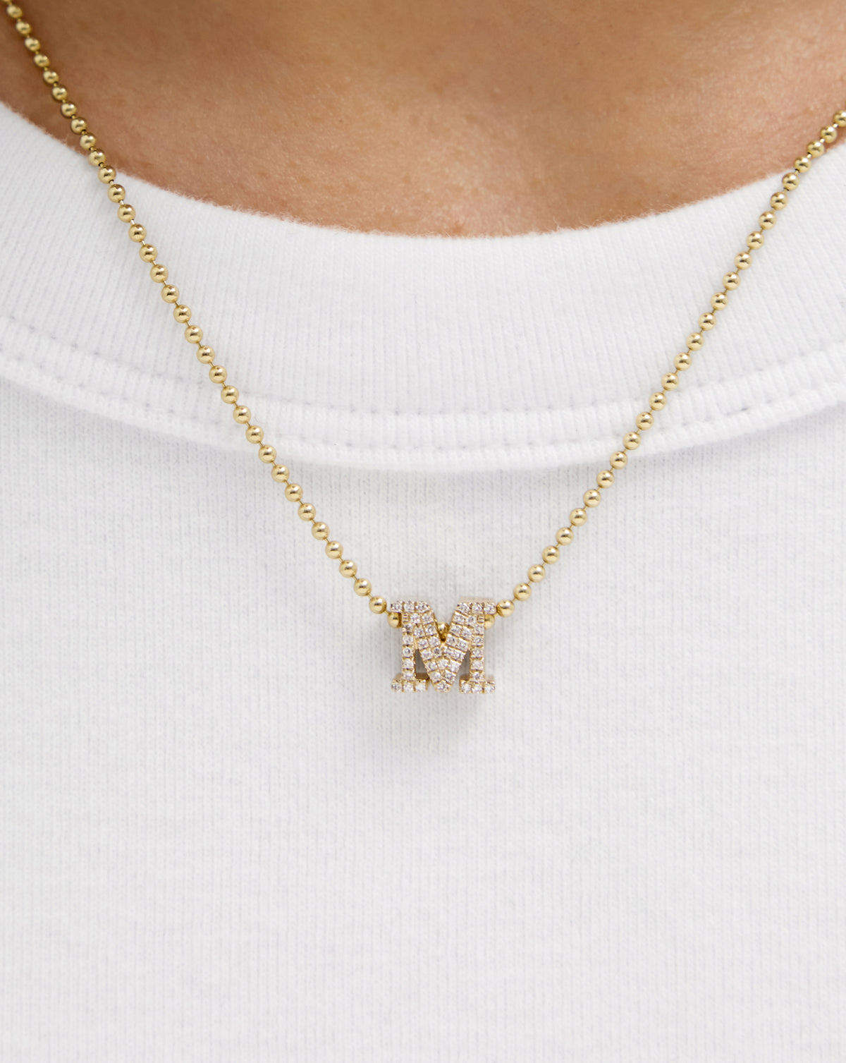 A close-up of a gold ball-chain necklace with a small pavé-set letter M pendant, worn on a model's neck over a white crewneck shirt. global