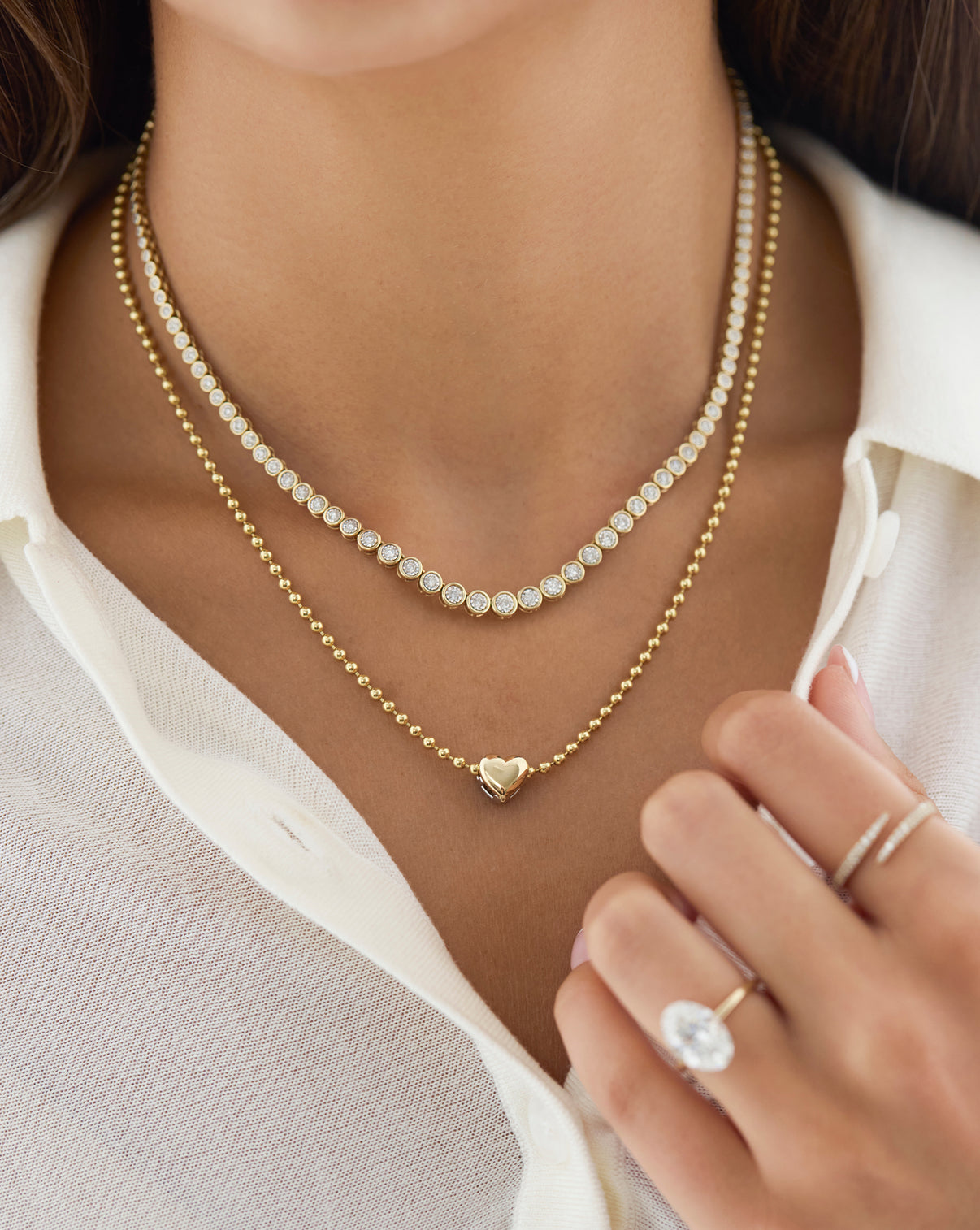 Close-up of a woman wearing layered necklaces, including a gold beaded chain with a small heart pendant and a delicate chain of clear stones, along with rings on her fingers.