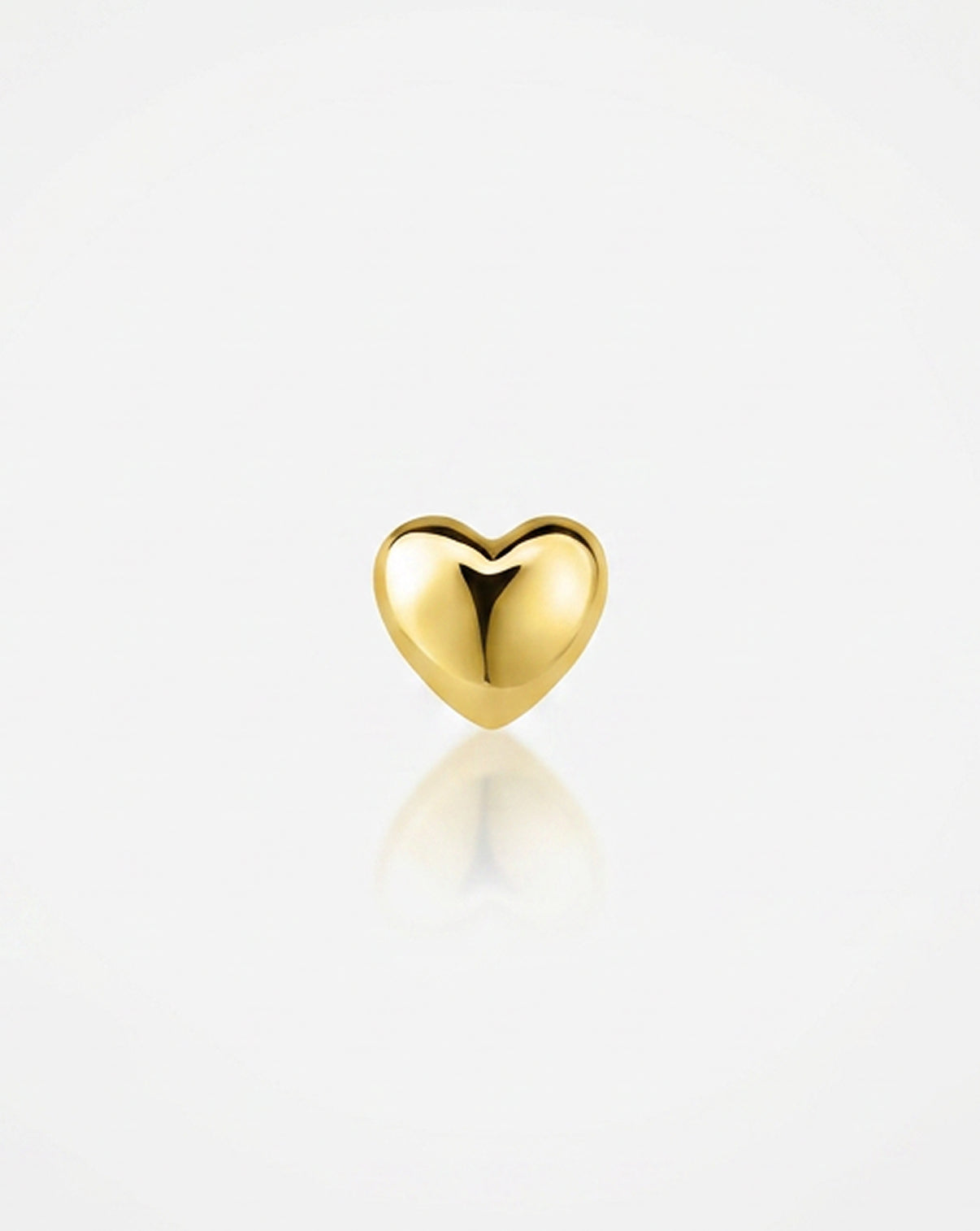 A small polished gold heart charm or pendant centered on a clean white background with a soft reflection below.