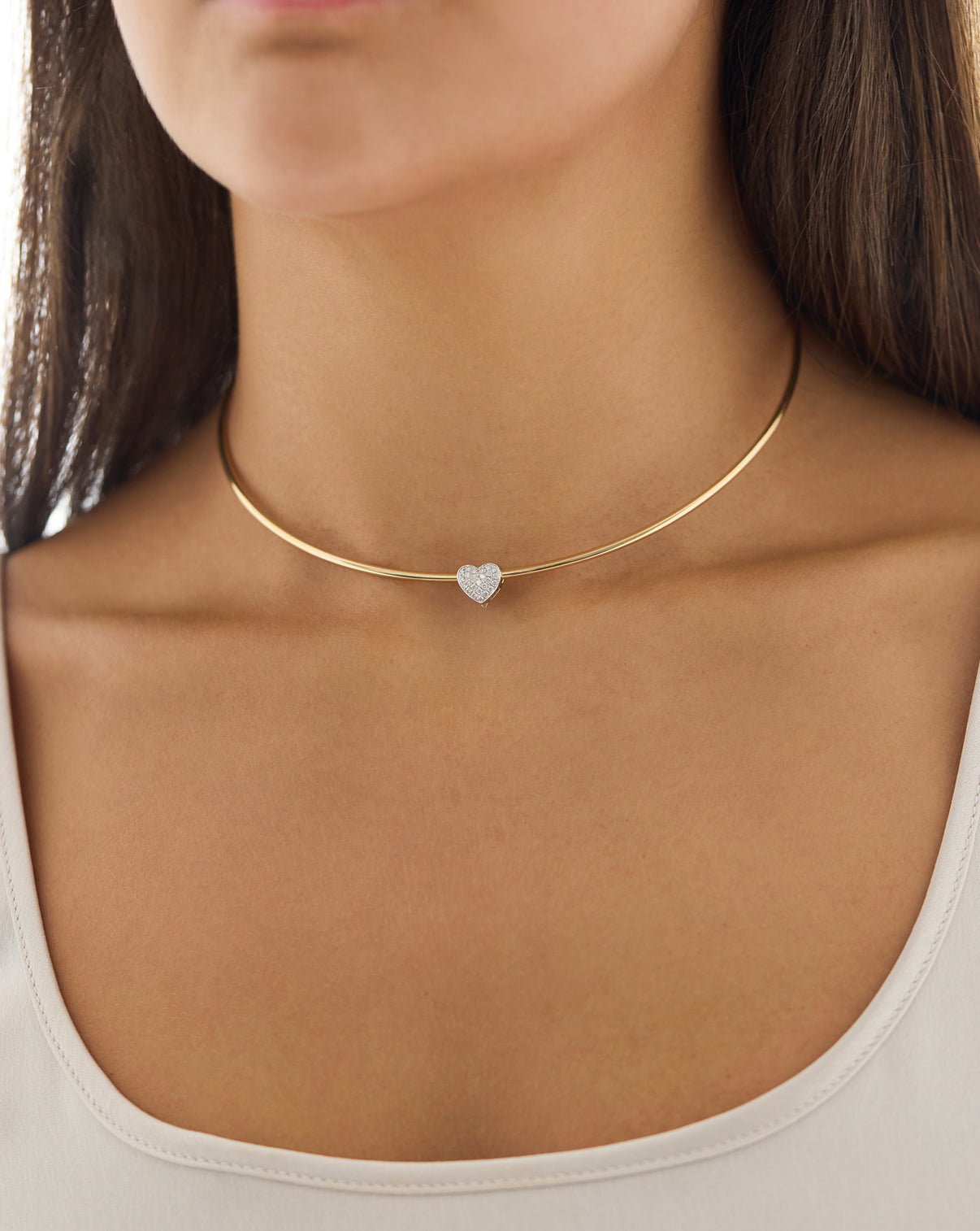 A close-up of a model wearing a delicate gold choker necklace with a small, heart-shaped clear gemstone at the center.