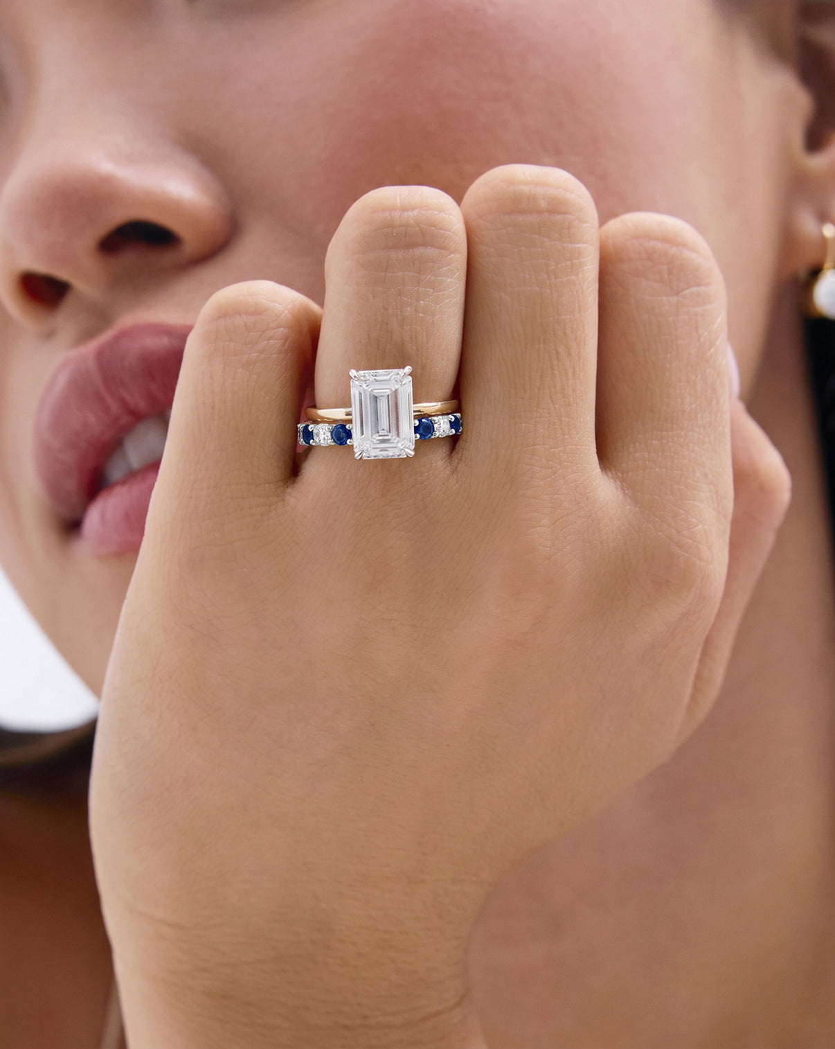 A close-up lifestyle shot of a model's hand wearing an emerald engagement ring with a classic sapphire and diamond wedding band