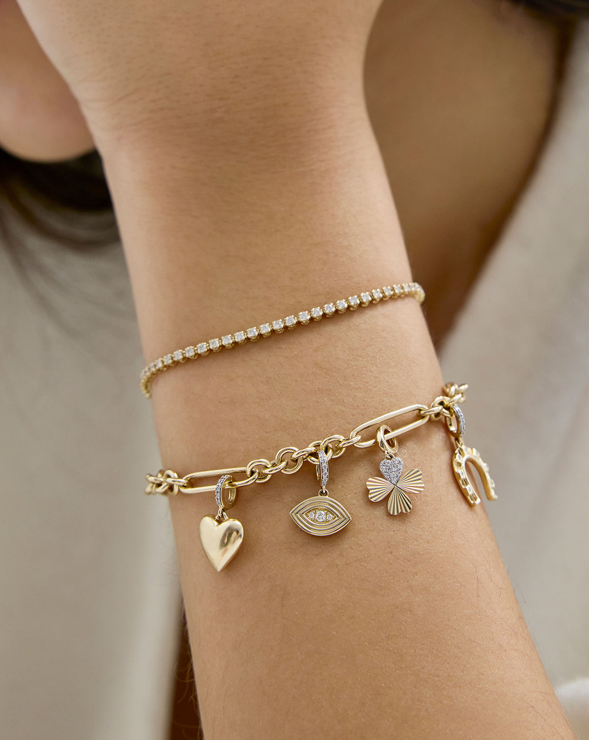 Close up of gold bracelet with a heart charm, an evil eye charm, a clover charm, and a horseshoe charm worn on model's wrist
