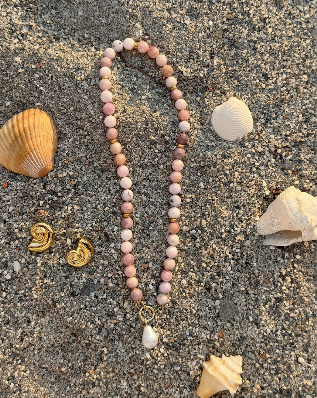 Pink Paloma Necklace laying in sand next our Nautilus Shell Earrings alongside a variety of seashells