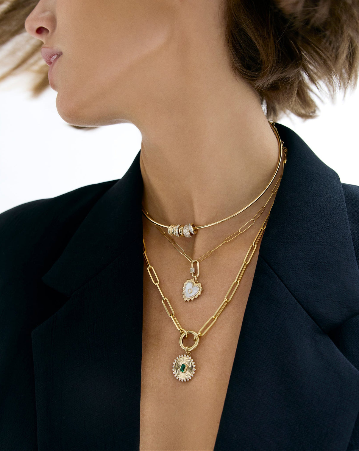 Close-up of a person wearing multiple gold necklaces with various pendants against a neutral background.