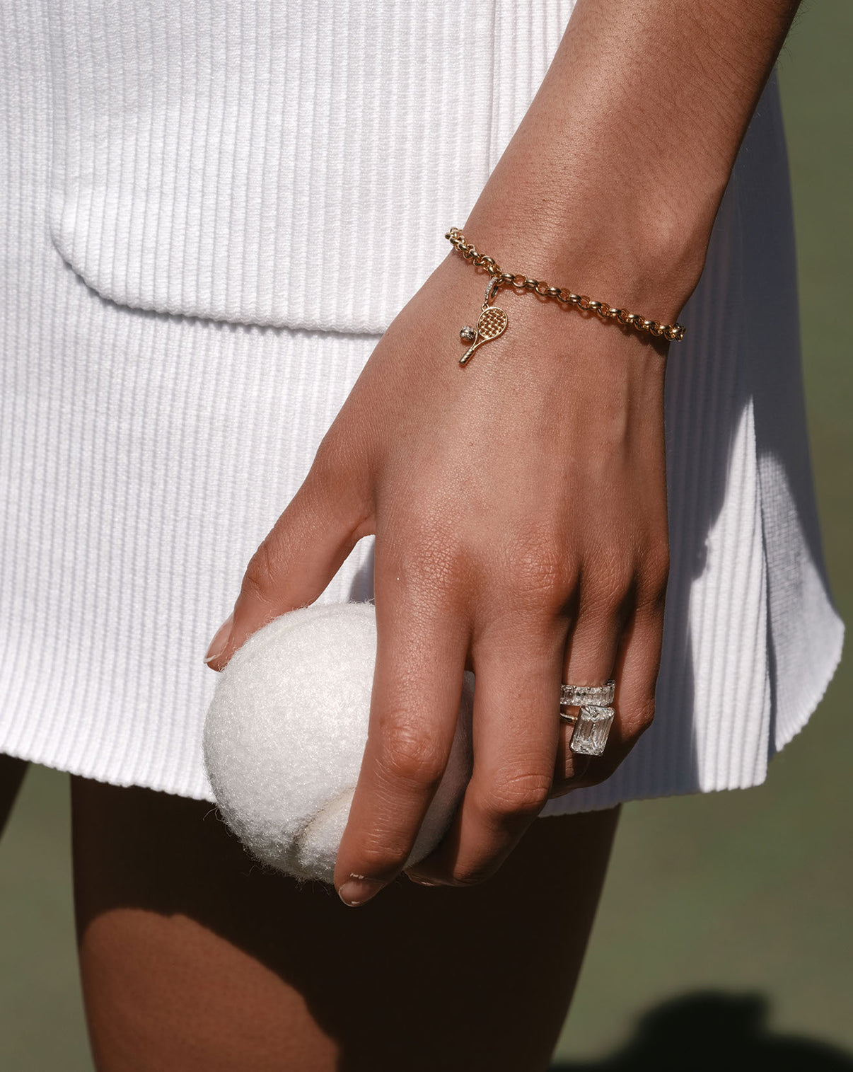 A close-up photo of a hand holding a white tennis ball. The model is wearing a yellow gold Hollow Rolo Chain Bracelet with a small tennis racket charm, and a diamond ring and band. 