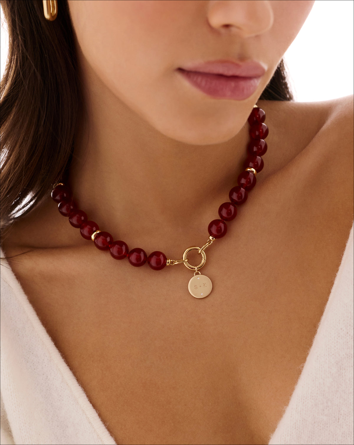 A close-up of a model wearing a dark red beaded necklace with a gold clasp and a round gold engravable charm.
