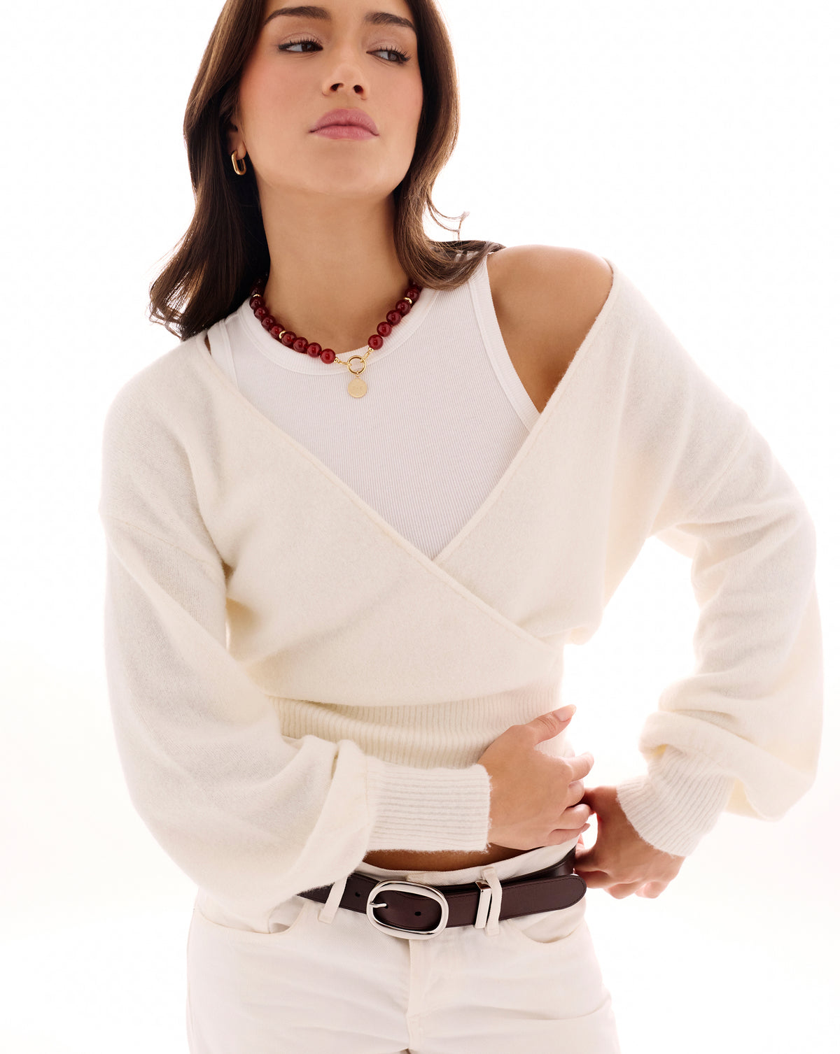 A woman wearing a cream knit wrap-style sweater over a white top, styled with a dark brown belt and a red beaded necklace, against a bright white background.