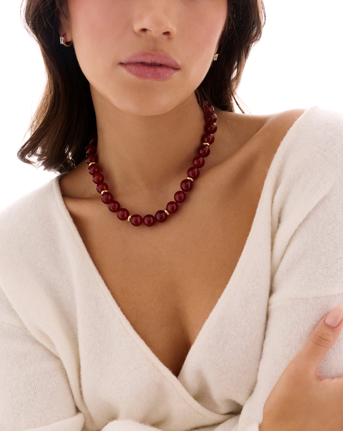 A close-up of a woman wearing a dark red beaded necklace with small gold spacer beads, styled with a cream knit wrap top against a bright white background.