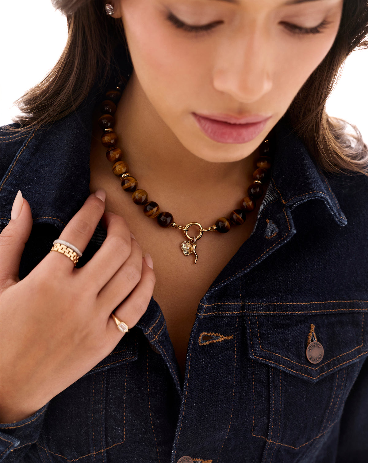 A close-up lifestyle shot of a woman wearing a tiger's eye beaded necklace with a gold heart charm, styled with a dark denim jacket.