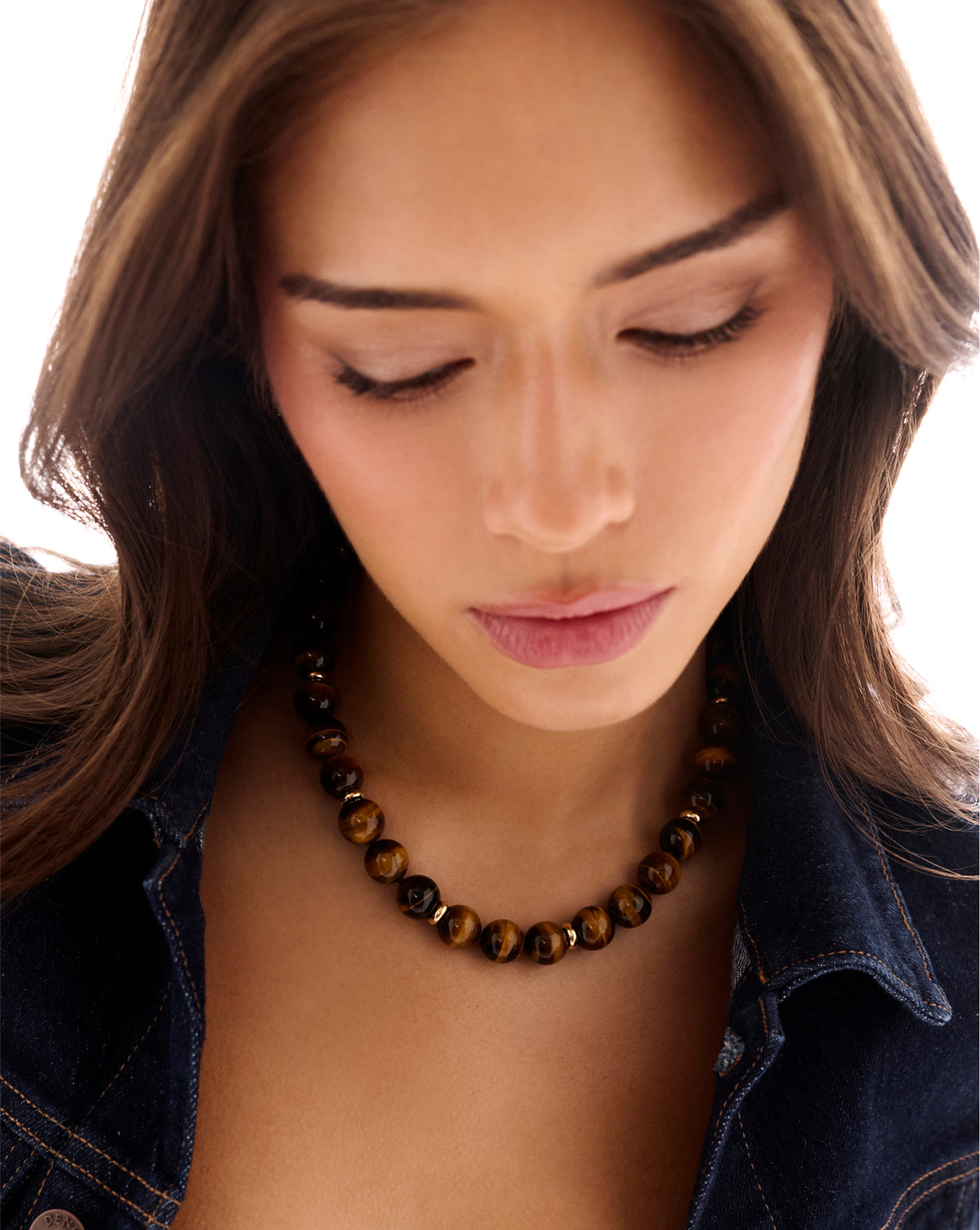 A close-up of a woman wearing a beaded necklace made of brown and black tiger's eye beads, paired with a dark denim jacket against a bright white background.