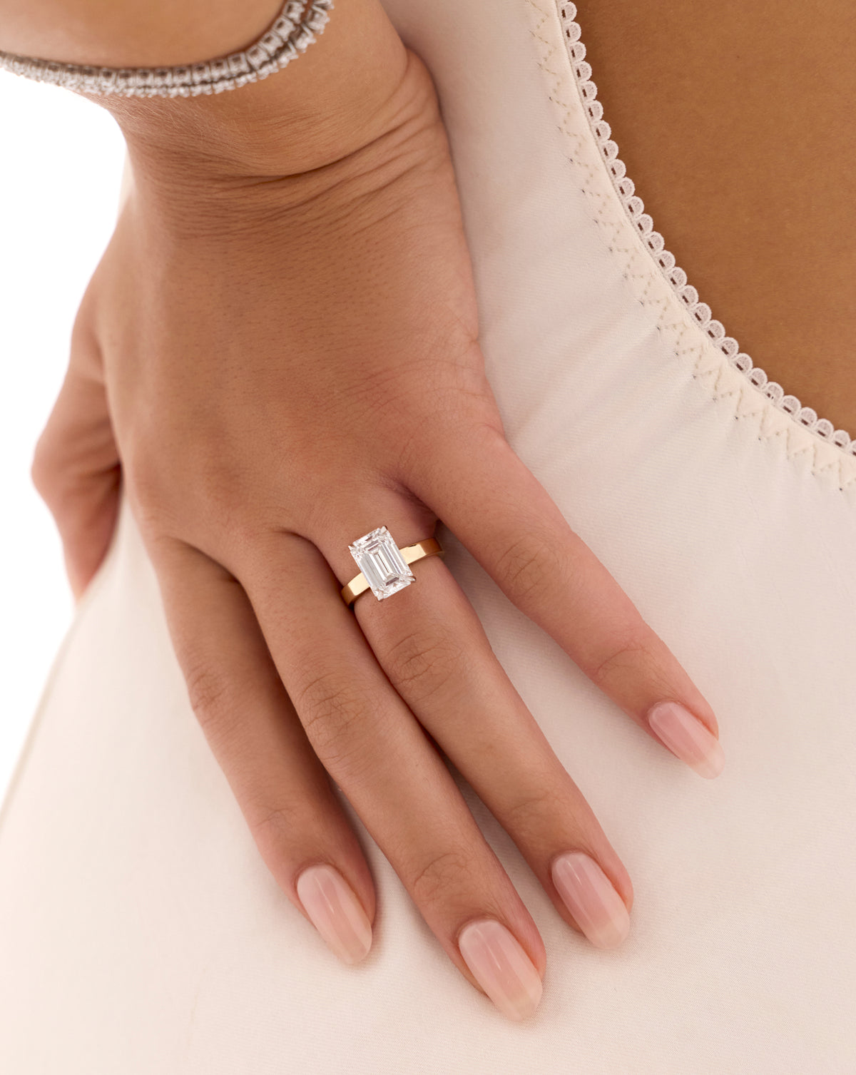 Close-up of a hand resting on ivory fabric, wearing a gold engagement ring with an emerald-cut diamond. 44830089281624 44830238638168 44830239326296