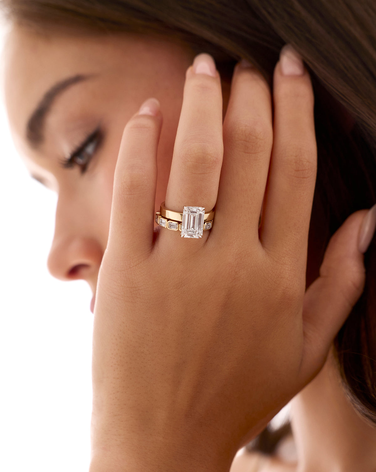 Model wearing 3.5 carat emerald cut lab-grown diamond engagement ring with cigar-style yellow gold band, hand held up against cheek showing ring close-up