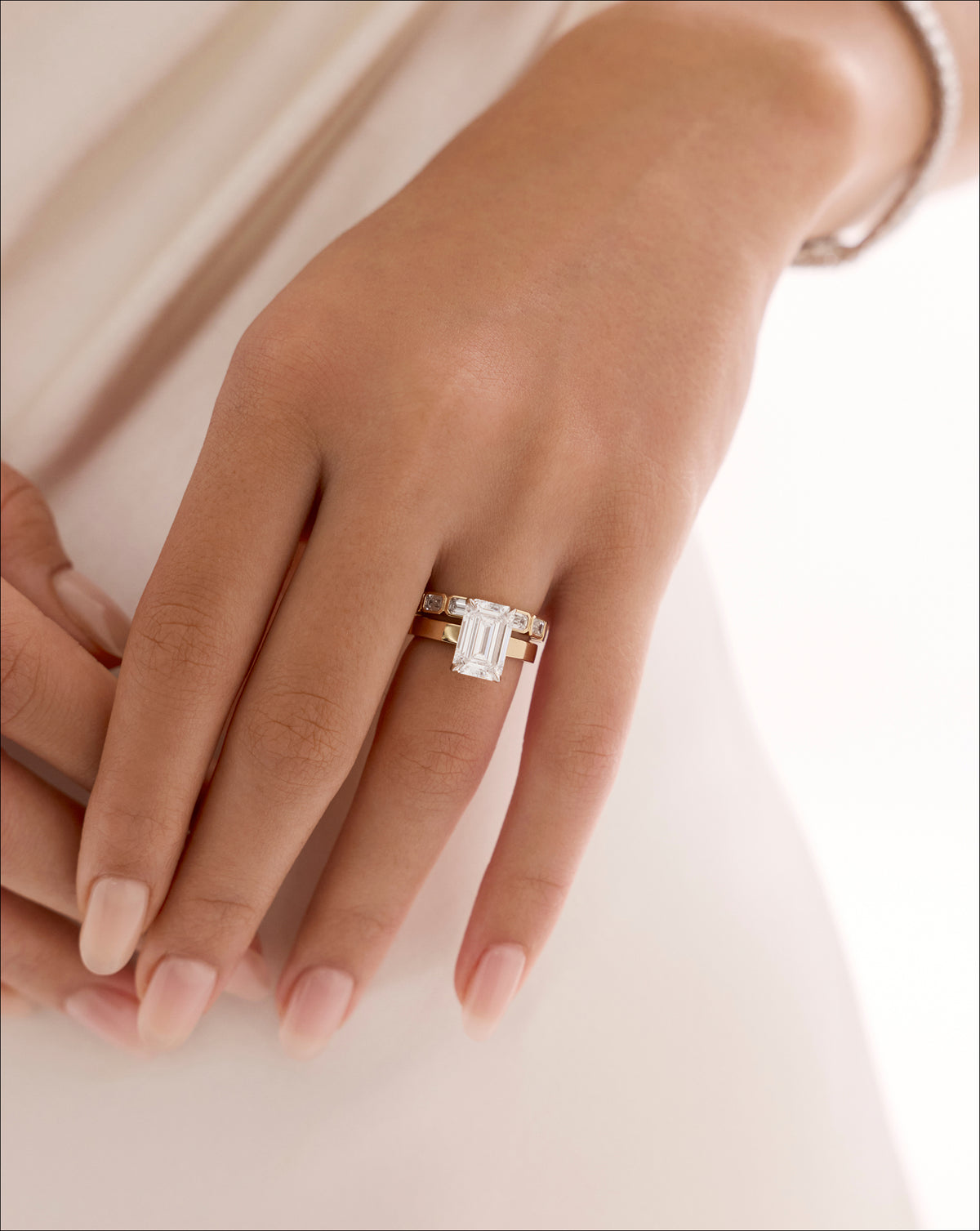 Close-up of a hand resting on fabric, wearing a gold engagement ring with an emerald-cut diamond and a matching pavé wedding band. 44830089281624 44830238638168 44830239326296