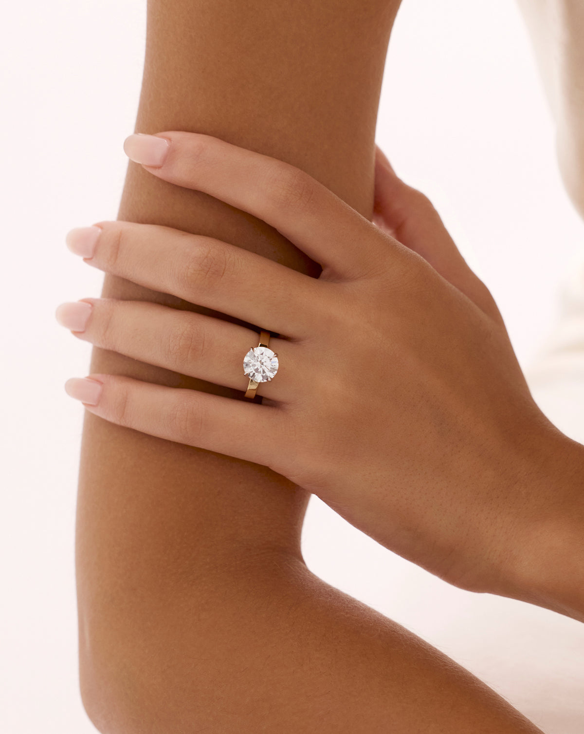 Close-up of a hand resting on the forearm, wearing a gold engagement ring with a round diamond in a cigar-style band. 44829996843096 44830238900312 44830239129688 