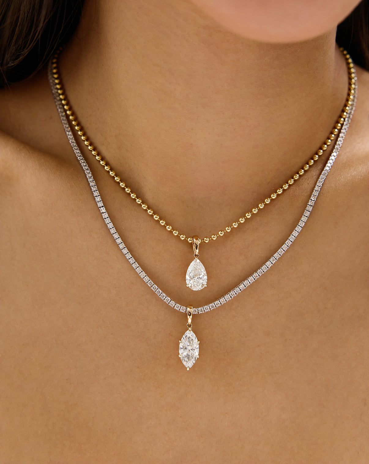 A close-up of a model's neck and chest wearing two layered necklaces: a gold beaded chain with a pear-cut pendant and a diamond tennis necklace with a marquise-cut pendant.