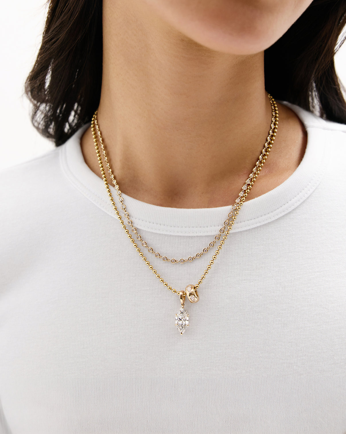 A close-up of a model wearing a double-layered gold necklace with a marquise diamond pendant, styled with a white crewneck t-shirt.
