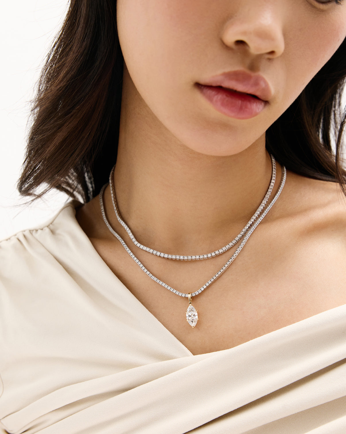 A close-up of a model wearing a double-layered tennis necklace with a marquise-cut pendant, paired with a cream-colored draped top.