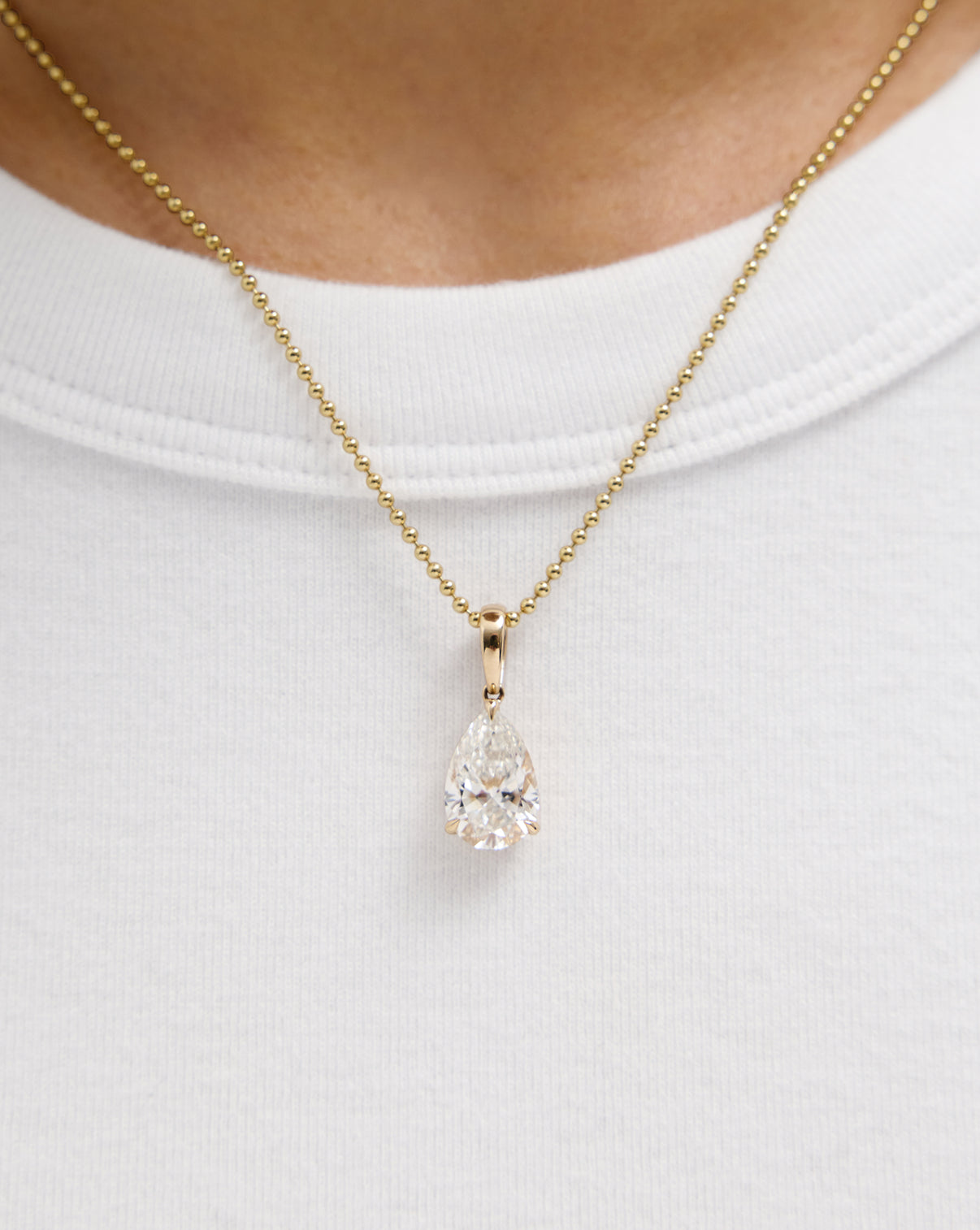 A gold ball-chain necklace with a pear-shaped diamond pendant, worn on a model's neck over a white t-shirt.
