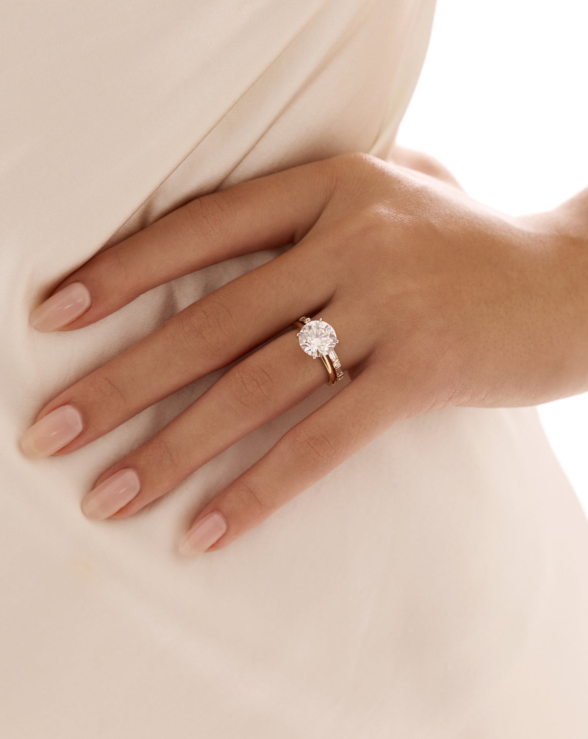 Close-up of a hand wearing the Baguette + Round Open Diamond Band in 14k yellow gold, stacked with a round diamond solitaire engagement ring against ivory satin. 14k Yellow Gold 14k Rose Gold 14k White Gold Platinum