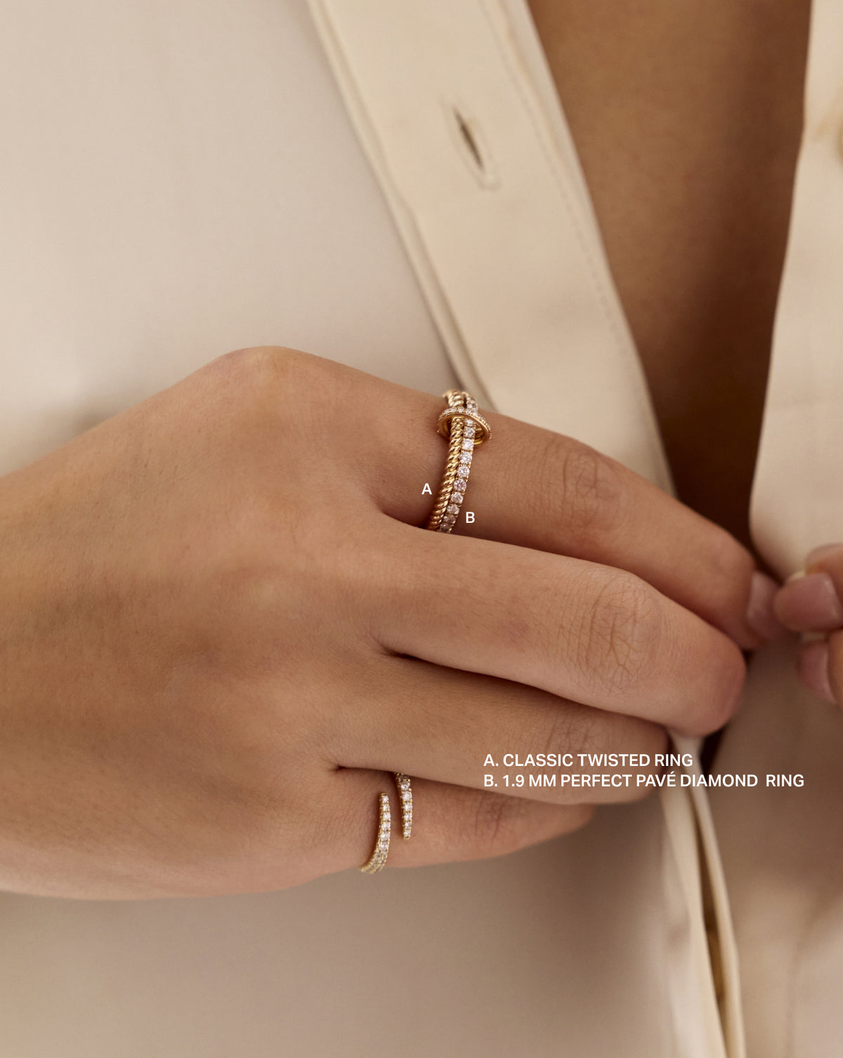 Close-up of a hand wearing the 14k yellow gold Classic Twisted Ring (A) and 1.9 mm Perfect Pavé Diamond Ring (B), connected by a Ring Clique Clasp while holding an ivory blouse. 14k yellow gold Sterling silver 