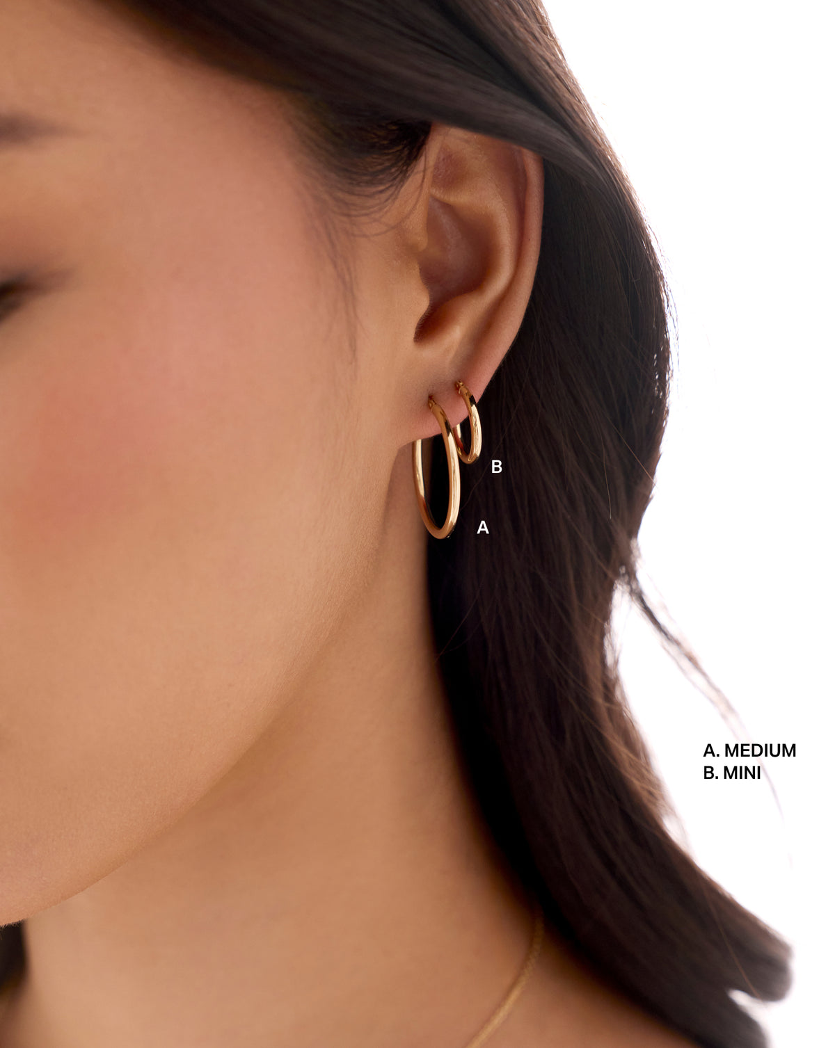 Close-up of a woman's ear wearing two gold hoop earrings, one medium-sized and one mini-sized, with text labels 'A. MEDIUM' and 'B. global