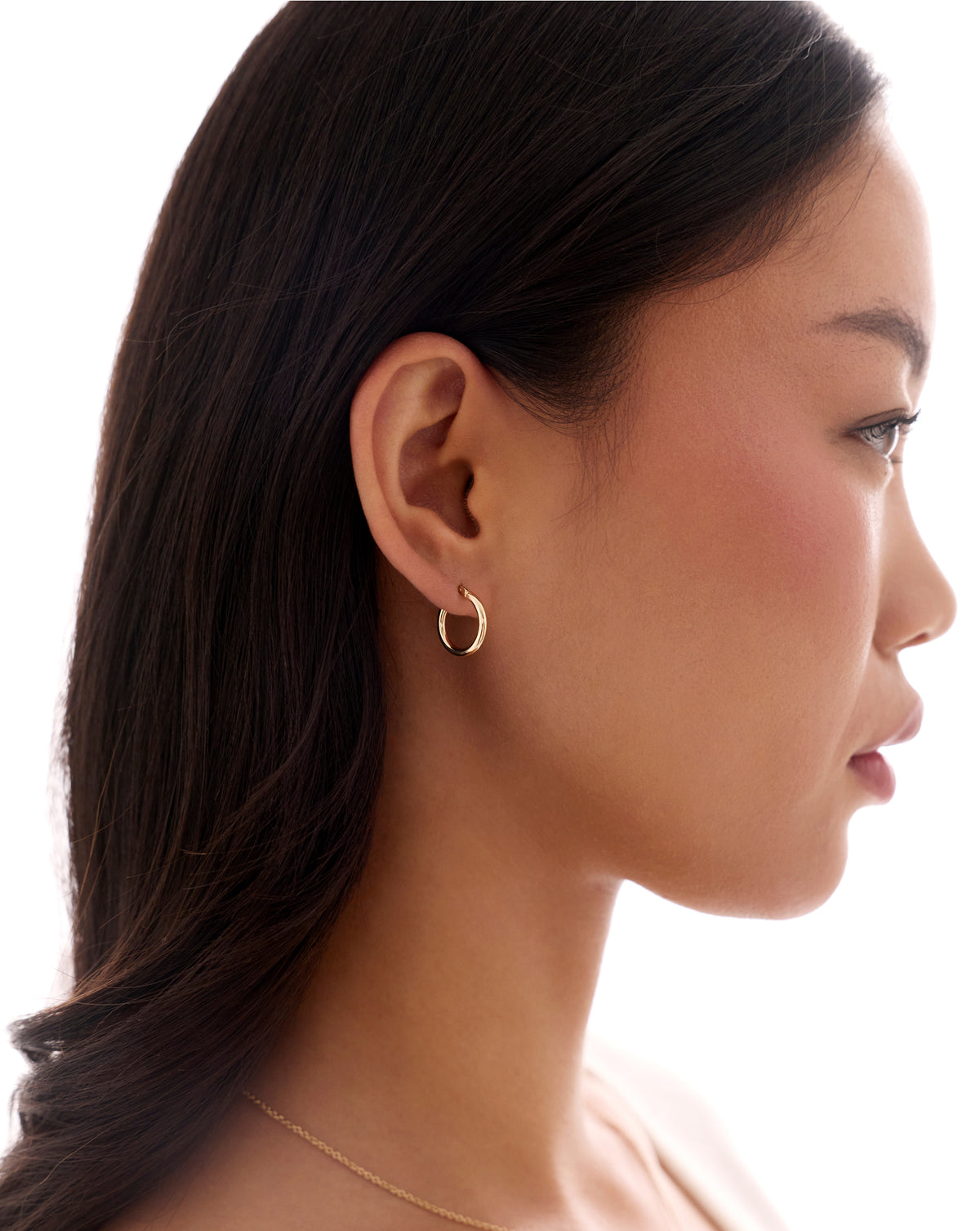 A close-up side profile of a woman with long dark hair wearing a delicate gold crescent hoop earring and a thin gold chain necklace against a bright white background. global