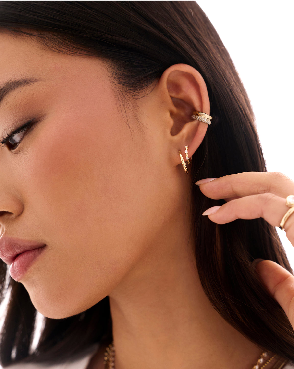 Close-up of a woman wearing a gold ear cuff with small hoop earrings, with her hand touching her ear against a clean white background. global