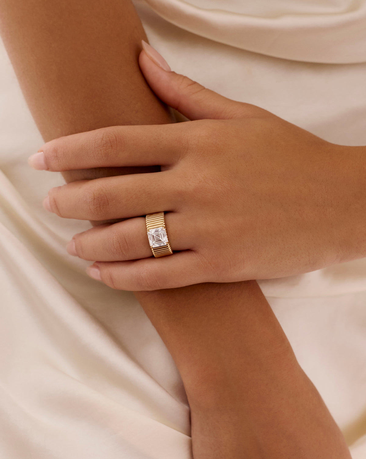 Hand wearing the Ribbon ring with a 2.85-carat Asscher-cut lab-grown diamond on a wide, fluted yellow gold band, resting on a forearm against ivory satin.