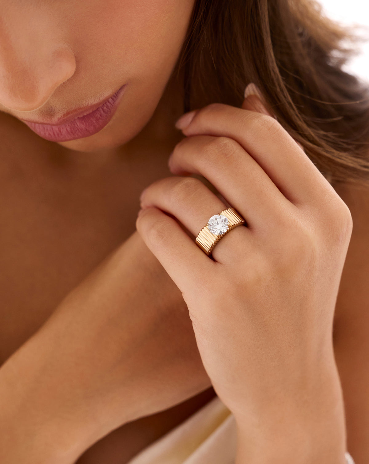 Close-up of a model wearing the Ribbon ring with a 2.00-carat round lab-grown diamond on a wide, fluted yellow gold band, hands clasped near her face.