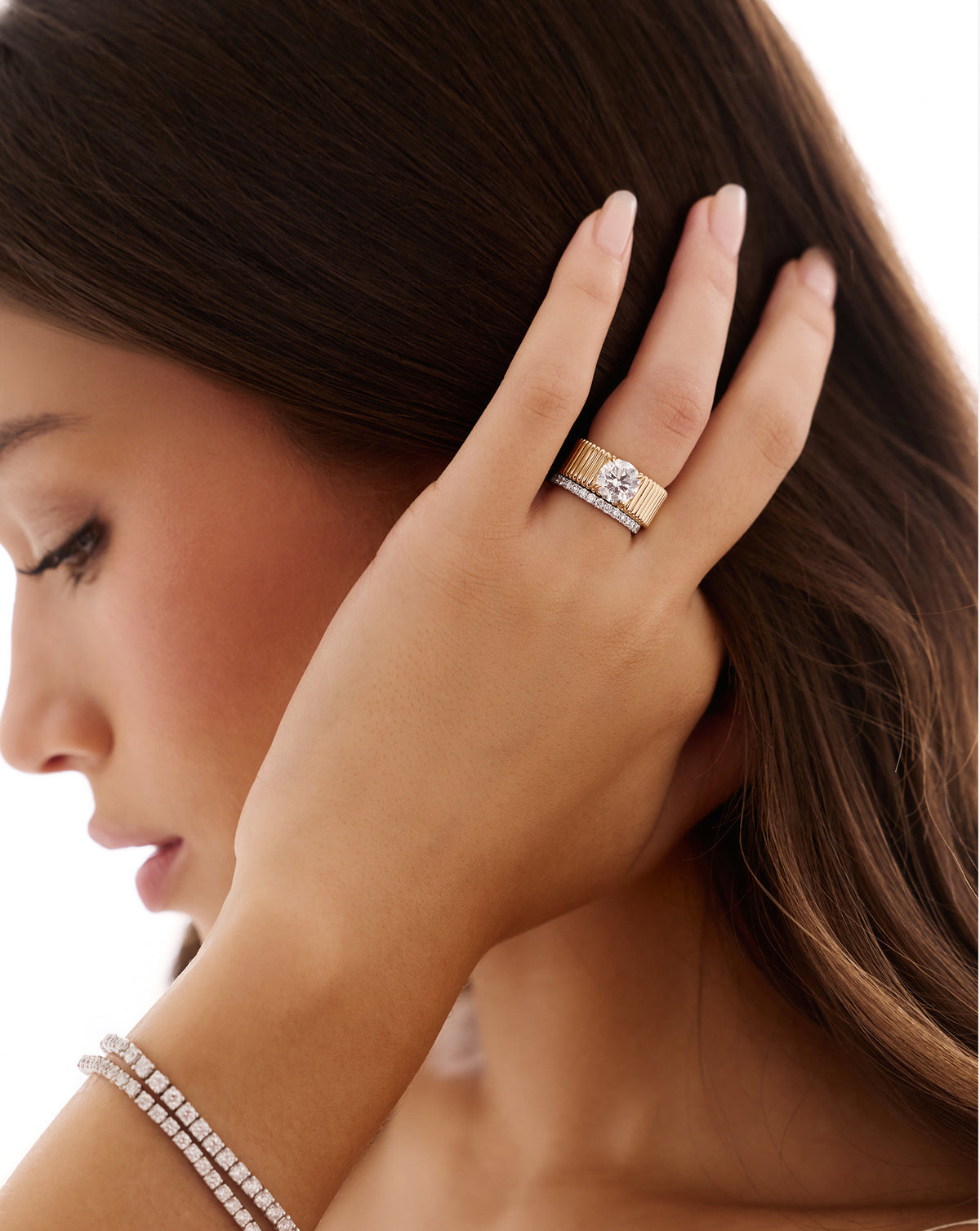 Model touching her hair while wearing the Ribbon ring with a 2.00-carat round lab-grown diamond on a wide, fluted yellow gold band, stacked with a slim diamond band.