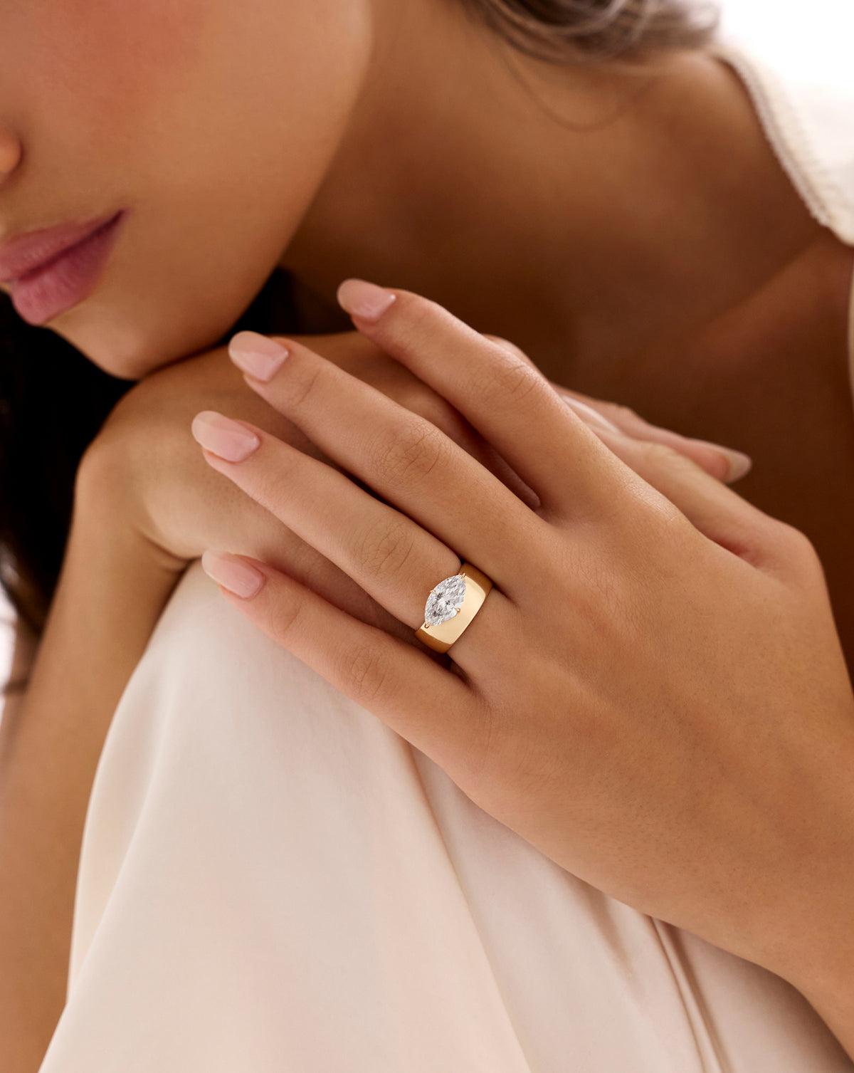 Close-up of a model wearing The Eclipse ring with a marquise-cut diamond on a wide 14K yellow gold half-dome band, resting her hand on ivory fabric. 67586767814744 67586638807128 67586768044120