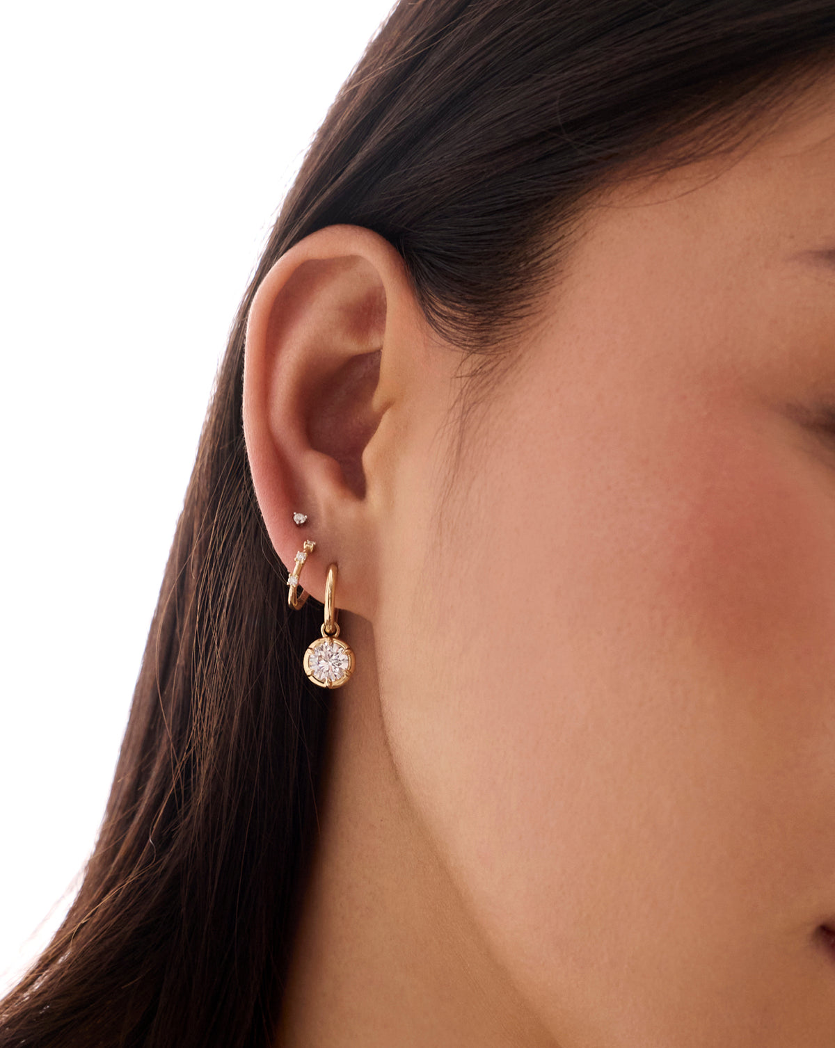Close-up profile of woman's ear showing a gold drop earring with a round diamond, worn alongside a second smaller stud earring in a higher piercing. global