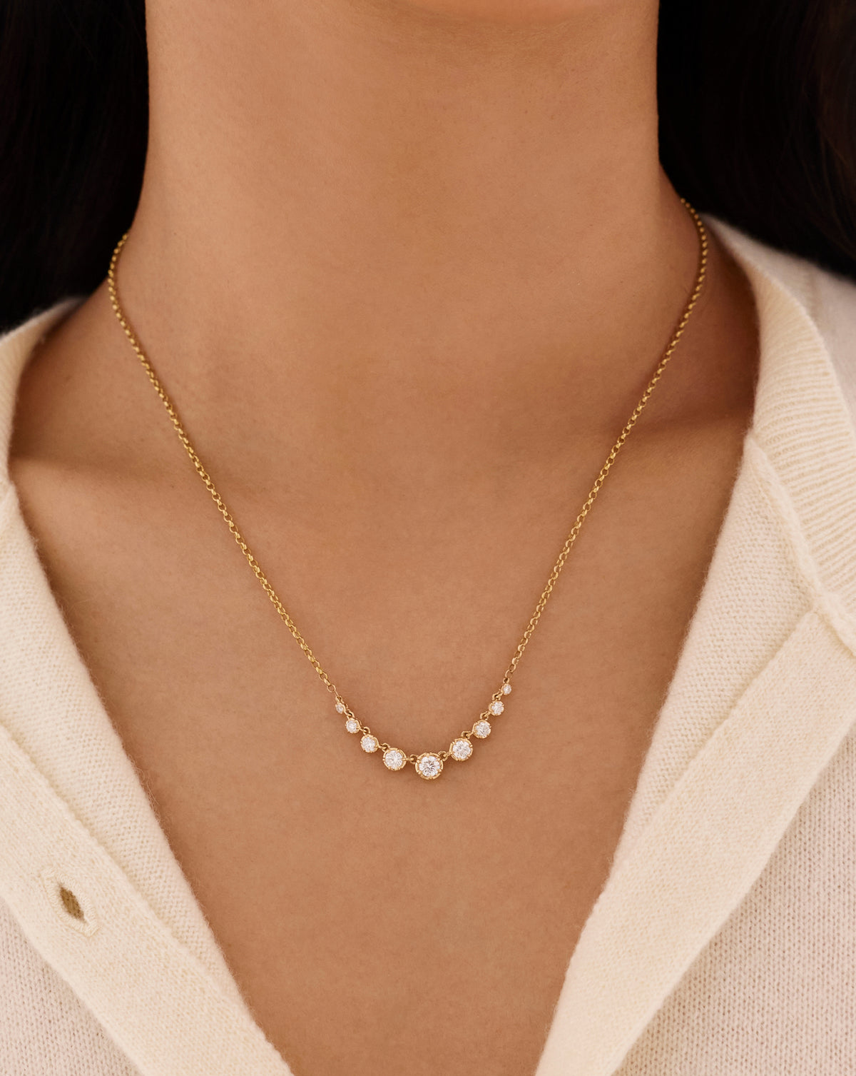 Close-up of a woman wearing a delicate gold chain necklace with a row of graduated round diamonds, styled with a cream knit cardigan. global