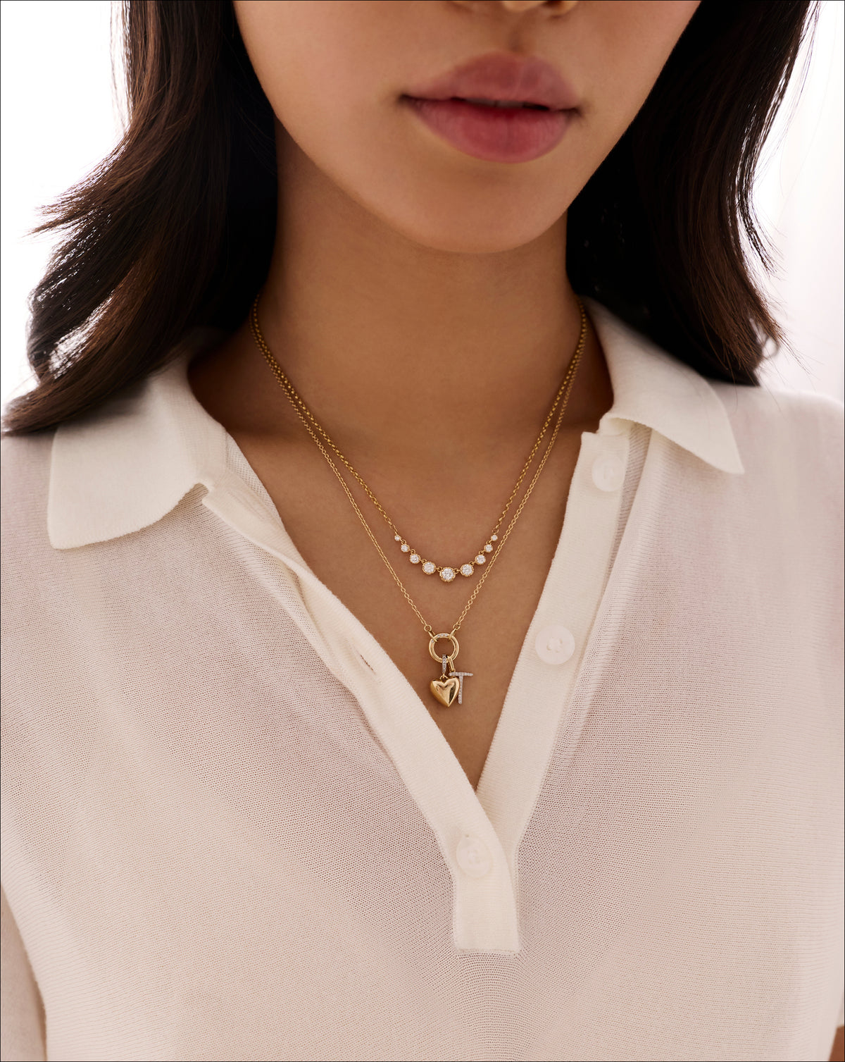 Close-up of woman's neckline wearing two layered gold necklaces — a graduated diamond chain and a second chain with a small heart charm — styled with a cream button-down top. global