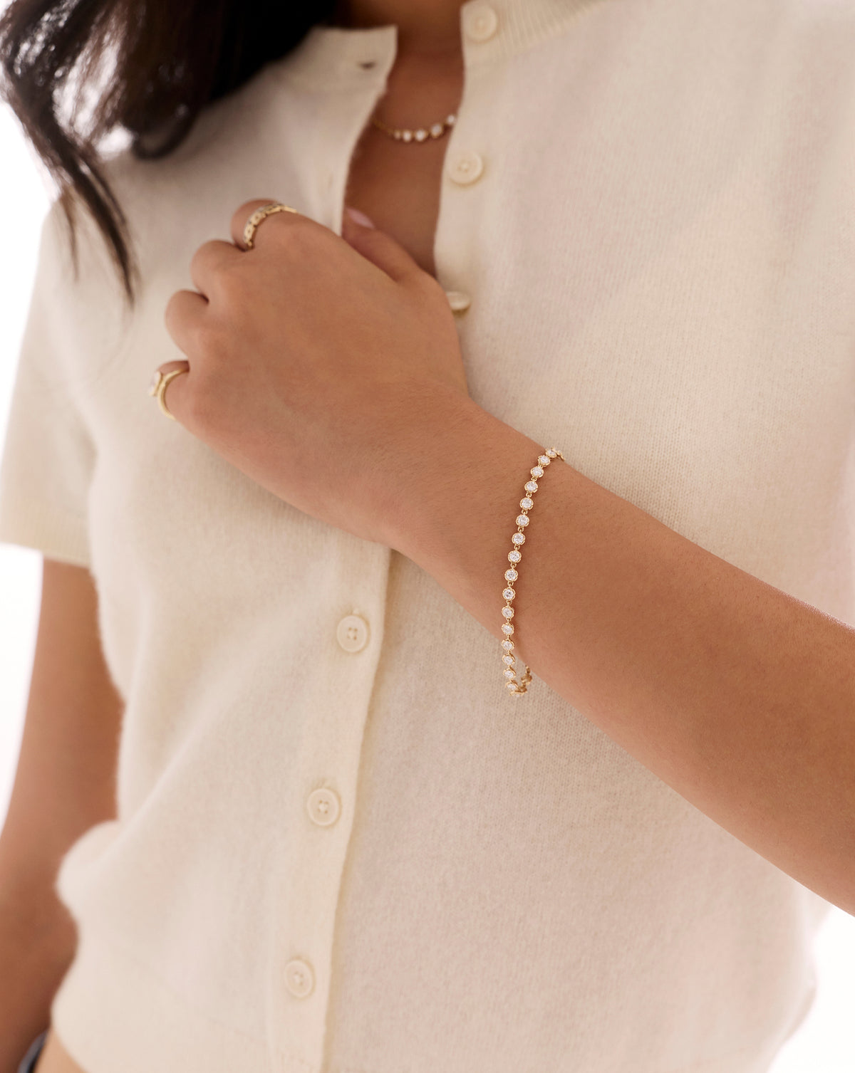 Gold tennis bracelet with sparkling round-cut stones worn on the wrist, styled with a white button-up top and gold rings. 44299697225816, 44299697586264