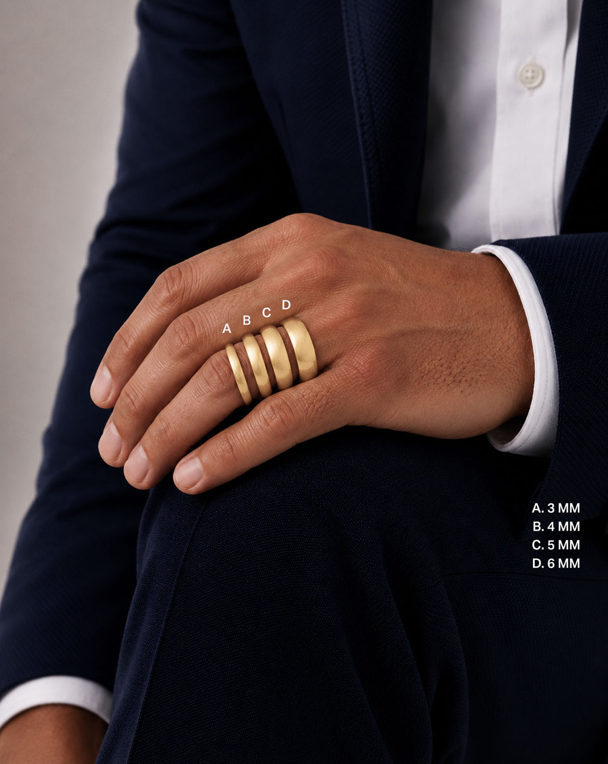 A close-up of a person's hand wearing four wide gold bands rings in varying widths (3mm, 4mm, 5mm, 6mm), styled with a dark navy blazer and white dress shirt. White text overlays indicate the ring widths. global