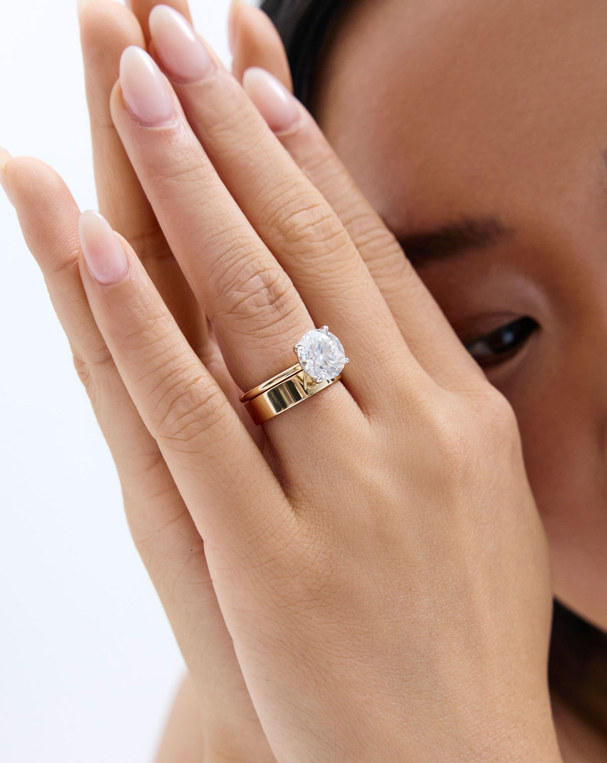 A close-up lifestyle shot of a woman's hand wearing a gold band ring with an engagement ring global
