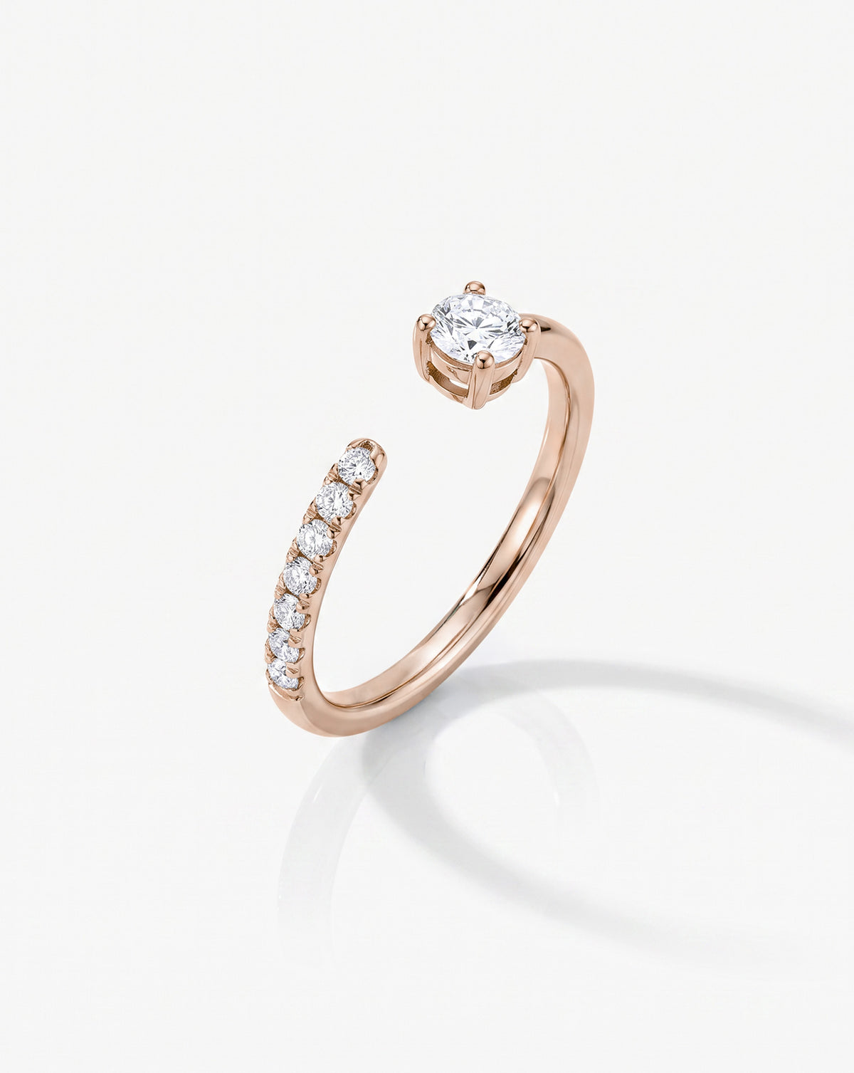 A 14k rose gold open band ring featuring a row of small round-cut diamondson one side and a larger center stone on the other, side view shown on a light gray background.