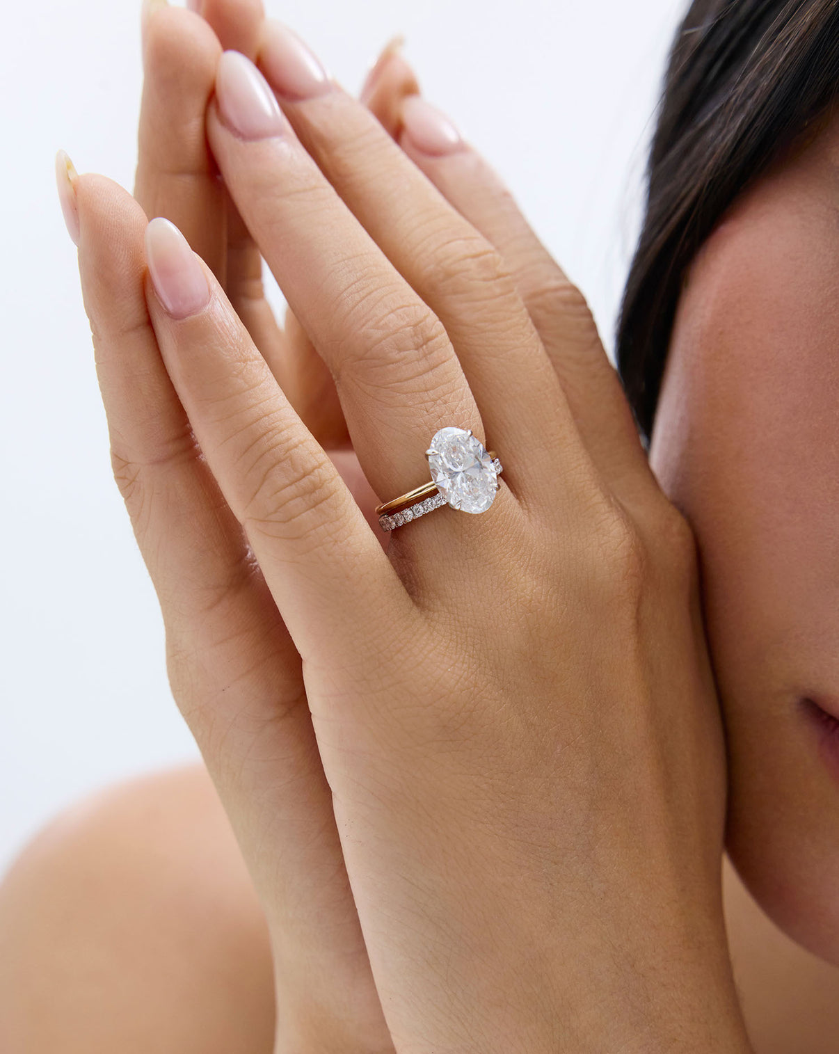 A close-up lifestyle shot of a woman's hands and face, wearing an oval engagement ring with a pavé band global.