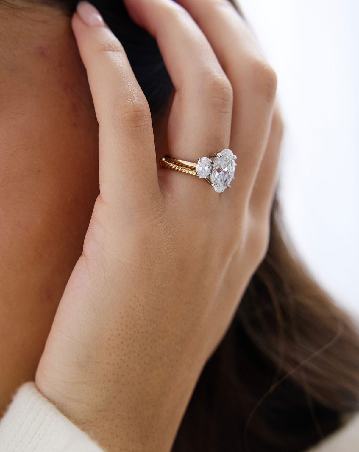 close up of model's hand wearing a 4 carat Oval Engagement Ring in 14k yellow gold Whisper Thin Three Stone with platinum prongs styled with a textured gold wedding band