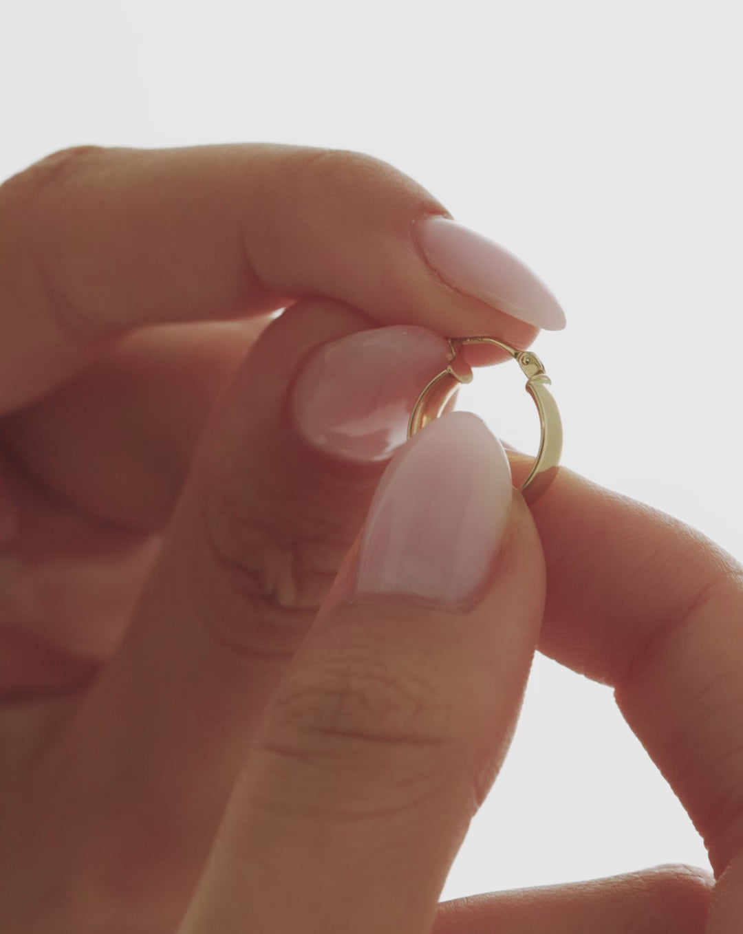 Close-up video of a 14k yellow gold wide mini hoop earring held between fingers, showing its rounded shape and hinged closure. global