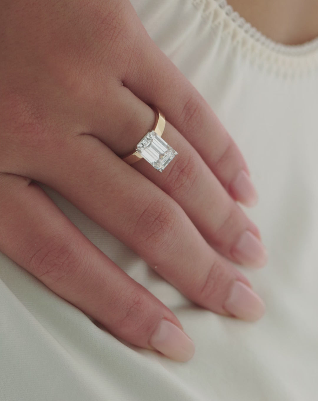 Video of a hand resting on ivory fabric, showcasing a gold engagement ring with an emerald-cut diamond in a wide flat band. 44830089281624 44830238638168 44830239326296