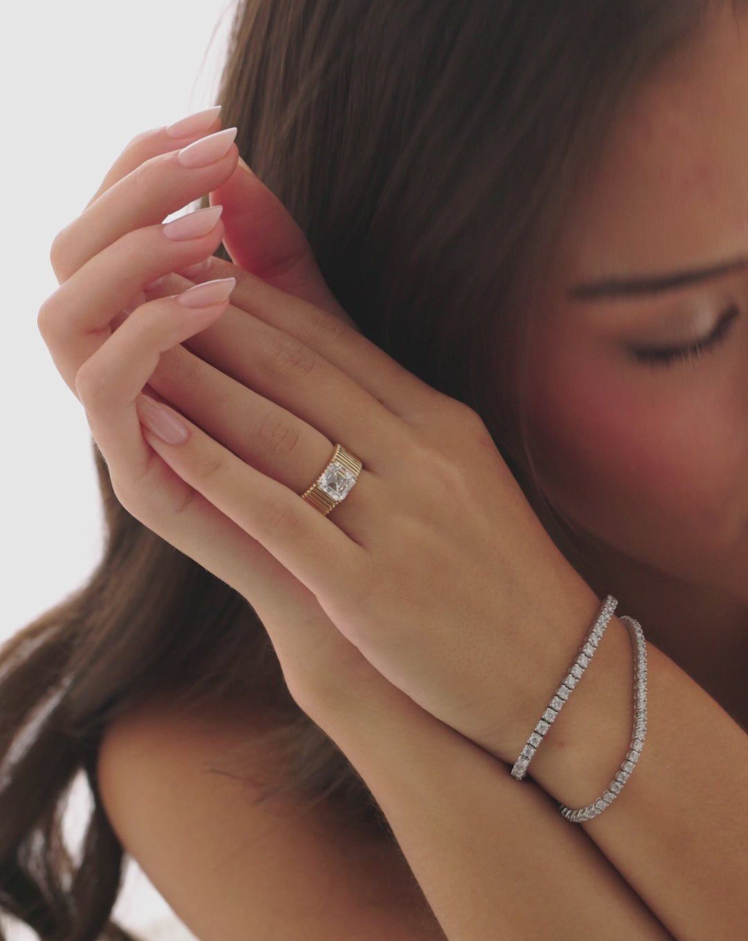 Video still of a model wearing the Ribbon ring with a 2.85-carat Asscher-cut lab-grown diamond on a wide, fluted yellow gold band, hands held beside her face.