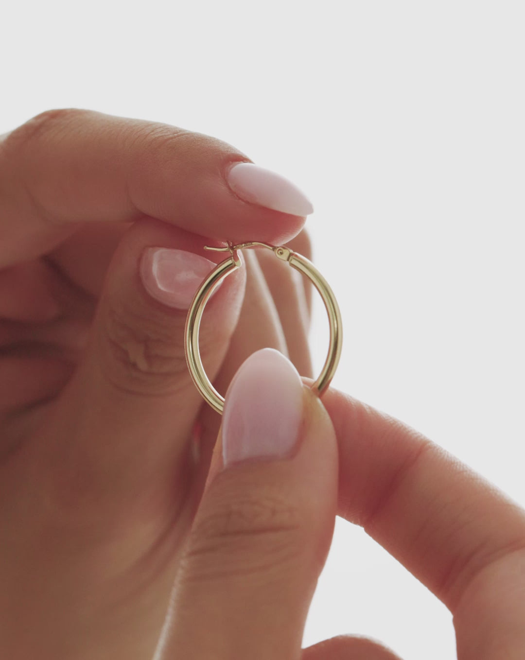 Video close-up of a 14k yellow gold essential medium hoop earring held between fingers, highlighting its classic rounded design and hinged closure. global