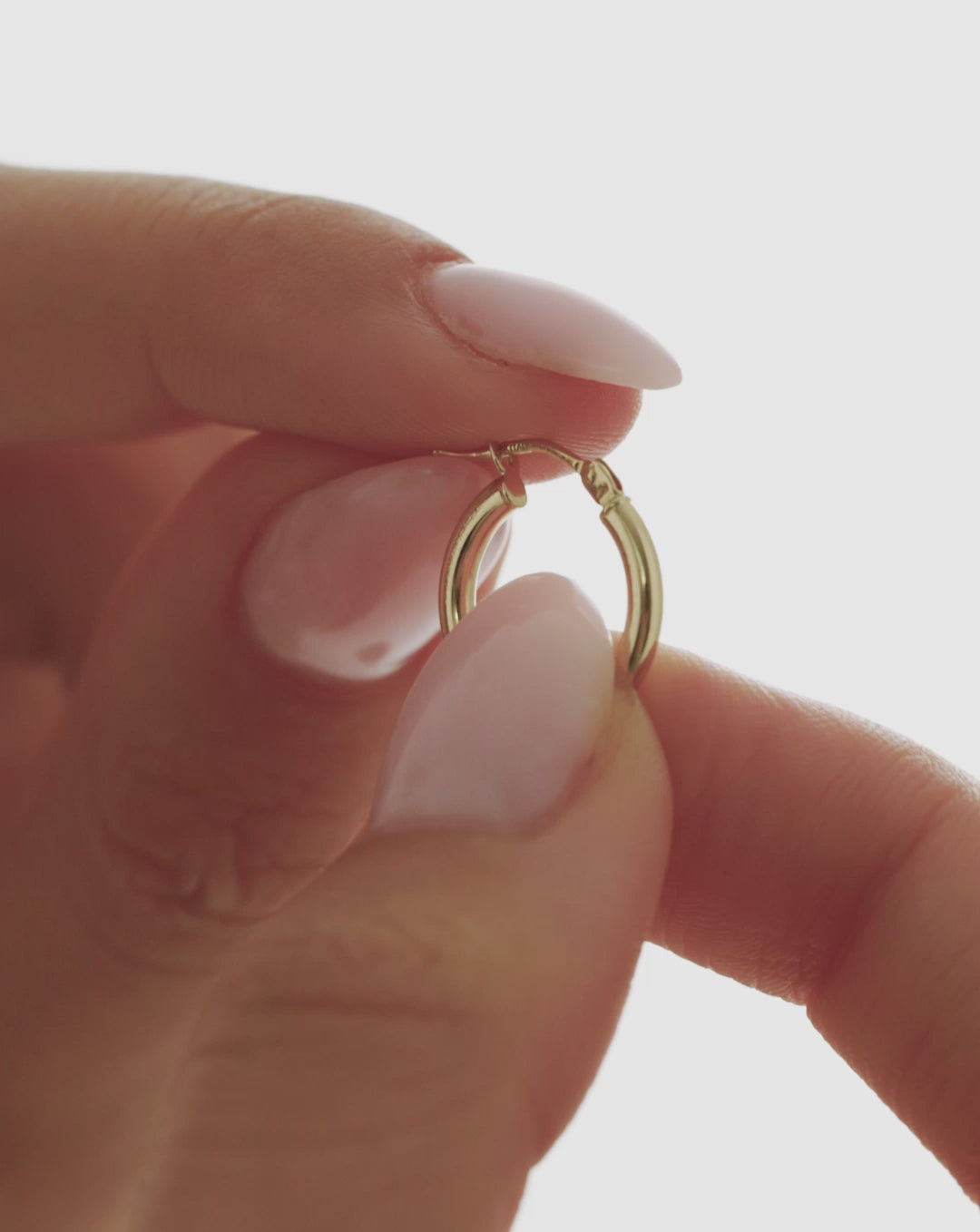 Video close-up of a 14k yellow gold essential mini hoop earring held between fingers, highlighting its classic rounded design and hinged closure. global